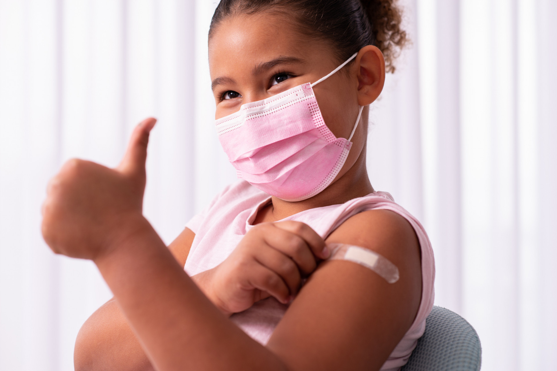 Child in mask showing thumb up after vaccination. There is a bandage on the child's arm.