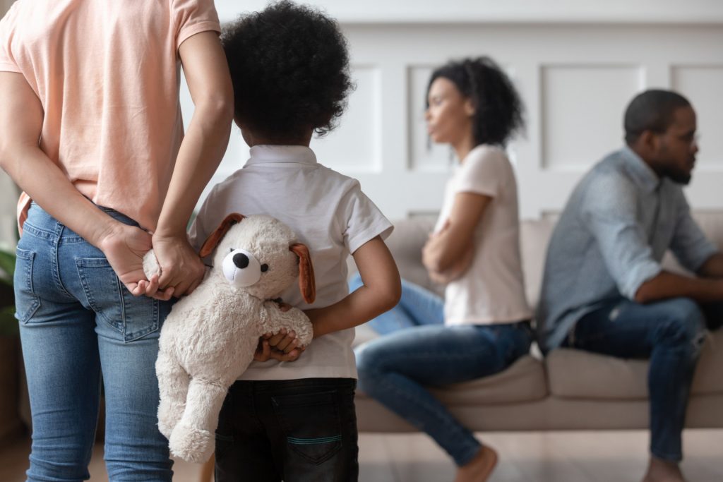 A child holds a stuffed animal toy behind their back and appears to look at two adults who are not facing each other.